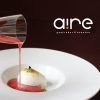 Un líquido rojo adorna con gracia un postre blanco en un plato, con el logotipo de Aire Gastrobar - Cocina mediterránea de autor en el fondo, resaltando la mezcla de técnicas tradicionales y vibrantes influencias mediterráneas. - ForTwo - Planes para parejas