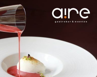 Un líquido rojo adorna con gracia un postre blanco en un plato, con el logotipo de Aire Gastrobar - Cocina mediterránea de autor en el fondo, resaltando la mezcla de técnicas tradicionales y vibrantes influencias mediterráneas. - ForTwo - Planes para parejas