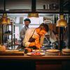 En la animada cocina del Restaurante María de la O, donde toma forma la “Cena romántica”, un chef emplata cuidadosamente los platos mientras sus compañeros charlan enérgicamente de fondo. - ForTwo - Planes para parejas