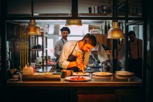 En la animada cocina del Restaurante María de la O, donde toma forma la “Cena romántica”, un chef emplata cuidadosamente los platos mientras sus compañeros charlan enérgicamente de fondo. - ForTwo - Planes para parejas
