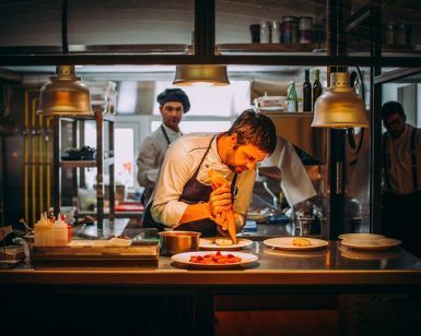 En la animada cocina del Restaurante María de la O, donde toma forma la “Cena romántica”, un chef emplata cuidadosamente los platos mientras sus compañeros charlan enérgicamente de fondo. - ForTwo - Planes para parejas
