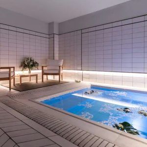 Experimente la serenidad en el moderno spa del Gran Hotel Inglés, con una piscina hundida y sillas de madera sobre paredes de azulejos blancos. La suave iluminación crea el ambiente perfecto para un relajante cóctel con acceso a jacuzzi y masaje en el Gran Hotel Inglés (Madrid). Disfrute de la tranquilidad. - ForTwo - Planes para parejas