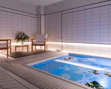 Experimente la serenidad en el moderno spa del Gran Hotel Inglés, con una piscina hundida y sillas de madera sobre paredes de azulejos blancos. La suave iluminación crea el ambiente perfecto para un relajante cóctel con acceso a jacuzzi y masaje en el Gran Hotel Inglés (Madrid). Disfrute de la tranquilidad. - ForTwo - Planes para parejas
