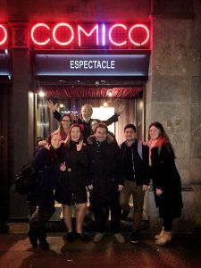 Un grupo de personas sonriendo y posando delante de un cartel iluminado de neón por la noche, listas para una velada de humor inolvidable en la bulliciosa entrada de Cómico Cómico en Barcelona para una Cena con espectáculo de humor. - ForTwo - Planes para parejas