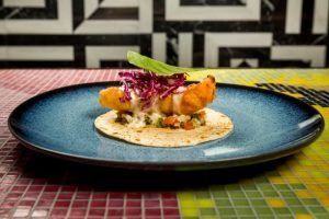 Taco de pescado frito con col morada y salsa en un plato azul en el Restaurante El Indiano Madrid-México, colocado sobre una mesa de mosaico colorido contra una pared geométrica. - ForTwo - Planes para parejas