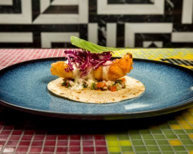 Taco de pescado frito con col morada y salsa en un plato azul en el Restaurante El Indiano Madrid-México, colocado sobre una mesa de mosaico colorido contra una pared geométrica. - ForTwo - Planes para parejas