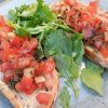 En el Restaurante Bon Gust (Valencia), disfruta de dos rebanadas de bruschetta con tomate picado y hierbas aromáticas, acompañadas de verduras frescas en un encantador plato azul. El sabor de Valencia te espera en cada bocado. - ForTwo - Planes para parejas