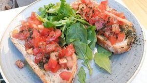 En el Restaurante Bon Gust (Valencia), disfruta de dos rebanadas de bruschetta con tomate picado y hierbas aromáticas, acompañadas de verduras frescas en un encantador plato azul. El sabor de Valencia te espera en cada bocado. - ForTwo - Planes para parejas
