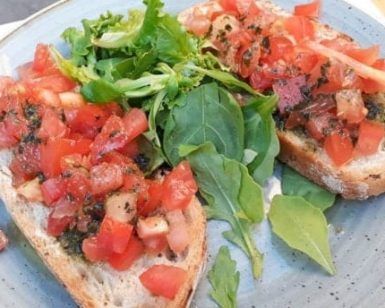 En el Restaurante Bon Gust (Valencia), disfruta de dos rebanadas de bruschetta con tomate picado y hierbas aromáticas, acompañadas de verduras frescas en un encantador plato azul. El sabor de Valencia te espera en cada bocado. - ForTwo - Planes para parejas