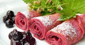 Crepes rosas espolvoreadas con azúcar glas, cubiertas con bayas y hojas en un plato blanco, reflejan la vitalidad del Taller de crepes de colores de Barcelona. - ForTwo - Planes para parejas