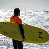 Alguien con un top naranja sostiene una tabla de surf amarilla, contemplando las olas del océano en Salobreña, un escenario perfecto para el "Curso de iniciación al surf de 2 horas en Salobreña". - ForTwo - Planes para parejas