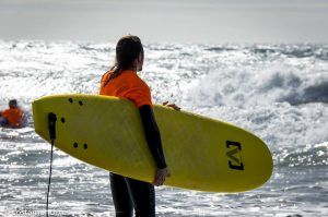 Alguien con un top naranja sostiene una tabla de surf amarilla, contemplando las olas del océano en Salobreña, un escenario perfecto para el "Curso de iniciación al surf de 2 horas en Salobreña". - ForTwo - Planes para parejas