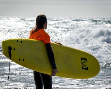 Alguien con un top naranja sostiene una tabla de surf amarilla, contemplando las olas del océano en Salobreña, un escenario perfecto para el "Curso de iniciación al surf de 2 horas en Salobreña". - ForTwo - Planes para parejas