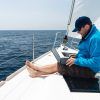 Un hombre con una chaqueta azul navega hábilmente con su computadora portátil en la cubierta de un velero en el vasto océano, posiblemente dominando las habilidades del Curso título patrón de embarcaciones de recreo PER con Cenautica (Madrid). - ForTwo - Planes para parejas