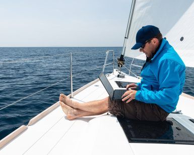 Un hombre con una chaqueta azul navega hábilmente con su computadora portátil en la cubierta de un velero en el vasto océano, posiblemente dominando las habilidades del Curso título patrón de embarcaciones de recreo PER con Cenautica (Madrid). - ForTwo - Planes para parejas