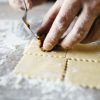 Primer plano de unas manos utilizando un tenedor para sellar el relleno dentro de láminas cuadradas de pasta fresca sobre una superficie enharinada, creando delicias culinarias como parte del Curso de pasta fresca en Madrid con degustación. - ForTwo - Planes para parejas