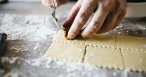 Primer plano de unas manos utilizando un tenedor para sellar el relleno dentro de láminas cuadradas de pasta fresca sobre una superficie enharinada, creando delicias culinarias como parte del Curso de pasta fresca en Madrid con degustación. - ForTwo - Planes para parejas