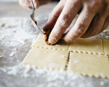 Primer plano de unas manos utilizando un tenedor para sellar el relleno dentro de láminas cuadradas de pasta fresca sobre una superficie enharinada, creando delicias culinarias como parte del Curso de pasta fresca en Madrid con degustación. - ForTwo - Planes para parejas