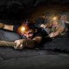Dos aventureros recorren un estrecho y oscuro túnel de cueva en una escena similar a "Darkness - Un juego de Maximum Escape Room (Barcelona)". Sus linternas frontales atraviesan la oscuridad mientras se apoyan en una cuerda, avanzando lentamente con una determinación que recuerda a un desafío de escape room. - ForTwo - Planes para parejas