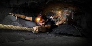 Dos aventureros recorren un estrecho y oscuro túnel de cueva en una escena similar a "Darkness - Un juego de Maximum Escape Room (Barcelona)". Sus linternas frontales atraviesan la oscuridad mientras se apoyan en una cuerda, avanzando lentamente con una determinación que recuerda a un desafío de escape room. - ForTwo - Planes para parejas