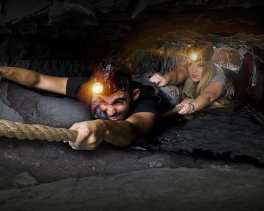 Dos aventureros recorren un estrecho y oscuro túnel de cueva en una escena similar a "Darkness - Un juego de Maximum Escape Room (Barcelona)". Sus linternas frontales atraviesan la oscuridad mientras se apoyan en una cuerda, avanzando lentamente con una determinación que recuerda a un desafío de escape room. - ForTwo - Planes para parejas