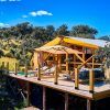 En Córdoba, entre árboles frondosos y un cielo azul brillante, encuentre tranquilidad en una terraza elevada de madera con sillones y un sofá. Una sombrilla proporciona sombra, haciéndose eco del encanto de "2 noches en Cabaña + cena + desayuno + decoración romántica en La Dehesa Experiences". - ForTwo - Planes para parejas