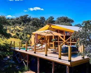 En Córdoba, entre árboles frondosos y un cielo azul brillante, encuentre tranquilidad en una terraza elevada de madera con sillones y un sofá. Una sombrilla proporciona sombra, haciéndose eco del encanto de "2 noches en Cabaña + cena + desayuno + decoración romántica en La Dehesa Experiences". - ForTwo - Planes para parejas