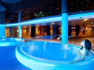 En el Hotel Diamante Beach, una mujer disfruta del moderno Circuito de Spa en Hotel Diamante Beach (Alicante) en una piscina cubierta iluminada en azul, bajo un techo estrellado que recuerda al sereno cielo nocturno de Alicante. - ForTwo - Planes para parejas