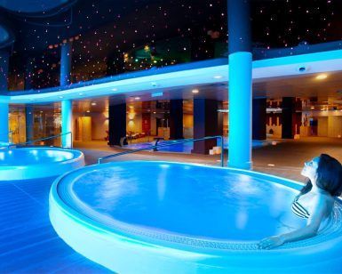 En el Hotel Diamante Beach, una mujer disfruta del moderno Circuito de Spa en Hotel Diamante Beach (Alicante) en una piscina cubierta iluminada en azul, bajo un techo estrellado que recuerda al sereno cielo nocturno de Alicante. - ForTwo - Planes para parejas