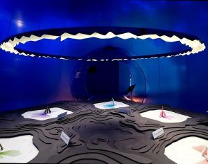 La sala azul futurista cuenta con una gran lámpara circular y exhibe el producto "Digital Impact" a través de su exhibición abstracta sobre una superficie texturizada. - ForTwo - Planes para parejas