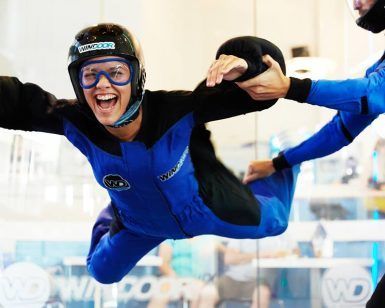 Una persona experimenta la emoción del paracaidismo indoor en Windoor, en Barcelona, vistiendo un traje azul y negro y sonriendo mientras un instructor le ayuda en el túnel de viento. - ForTwo - Planes para parejas