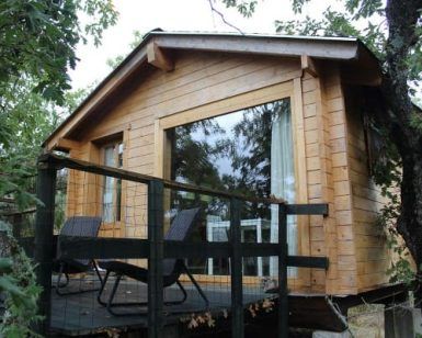Ubicada en la exuberante vegetación de Cáceres, la Cabaña Musgo en Dormir en los Árboles ofrece un refugio tranquilo. La cabaña de madera se asienta sobre una plataforma con amplios ventanales y dos sillas negras en la terraza, ofreciendo un refugio idílico. - ForTwo - Planes para parejas