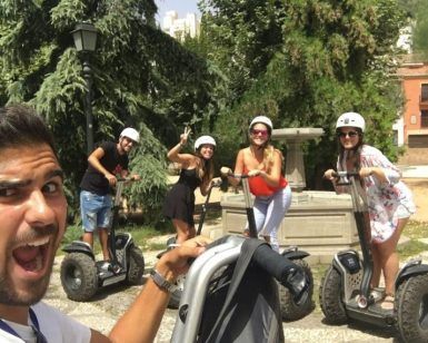 Cuatro personas en Segways, con cascos y sonrientes, posan en un parque con árboles y edificios mientras disfrutan de la pintoresca "Hacer una ruta en Segway por el Albaicín" a través del pintoresco paisaje. - ForTwo - Planes para parejas