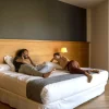 Dos huéspedes se relajan y conversan en una moderna habitación del Hotel Món, con suelos de madera y un gran ventanal, disfrutando de la serenidad del cercano Monasterio de Sant Benet. Disfrute de su estancia en el Hotel Món 4 y sumérjase en una experiencia medieval en el Monasterio de Sant Benet (Bages, Barcelona). - ForTwo - Planes para parejas