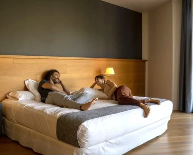 Dos huéspedes se relajan y conversan en una moderna habitación del Hotel Món, con suelos de madera y un gran ventanal, disfrutando de la serenidad del cercano Monasterio de Sant Benet. Disfrute de su estancia en el Hotel Món 4 y sumérjase en una experiencia medieval en el Monasterio de Sant Benet (Bages, Barcelona). - ForTwo - Planes para parejas