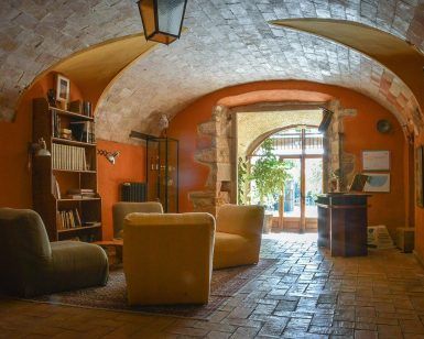 Una acogedora sala que evoca el encanto de una masía catalana del siglo XVIII, con techo abovedado, cómodos sillones y estanterías. La entrada, soleada y llena de plantas, da una cálida bienvenida, como la Escapada de quesos de Mas Alba. - ForTwo - Planes para parejas