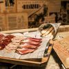 Disfruta de una tabla de embutidos en l'Espai Quera, Barcelona, con productos locales: carnes cortadas en papel de periódico, palitos de pan en un cono y una bolsa marrón sobre una mesa de cristal. - ForTwo - Planes para parejas