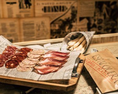 Disfruta de una tabla de embutidos en l'Espai Quera, Barcelona, con productos locales: carnes cortadas en papel de periódico, palitos de pan en un cono y una bolsa marrón sobre una mesa de cristal. - ForTwo - Planes para parejas