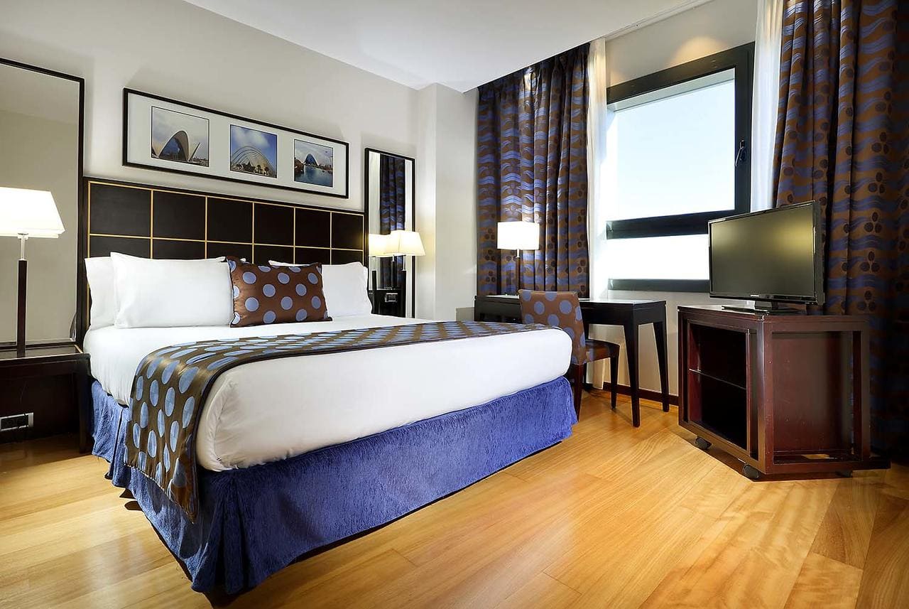 Experimente una comodidad inigualable en nuestra habitación bellamente decorada del Hotel Eurostars Gran Valencia, que cuenta con una cama doble con almohadas estampadas, pisos de madera, escritorio, TV y arte enmarcado en la pared. - ForTwo - Planes para parejas