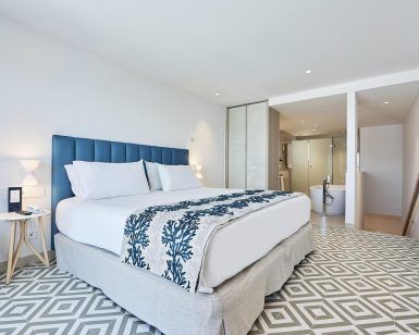 Disfrute de la elegancia moderna en el Hotel Eurostars Ibiza, donde le esperan una cama espaciosa, detalles azules, azulejos geométricos y un baño abierto con bañera y ducha. - ForTwo - Planes para parejas