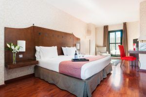 Descubra el encanto del Hotel Exe Agora en Cáceres, que ofrece una acogedora habitación con una cama amplia, detalles en rosa y suelo de madera. Relájese en el sillón rojo junto a la ventana, disfrutando de flores y vino servidos en una bandeja. - ForTwo - Planes para parejas