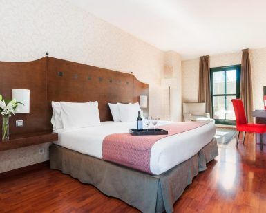 Descubra el encanto del Hotel Exe Agora en Cáceres, que ofrece una acogedora habitación con una cama amplia, detalles en rosa y suelo de madera. Relájese en el sillón rojo junto a la ventana, disfrutando de flores y vino servidos en una bandeja. - ForTwo - Planes para parejas