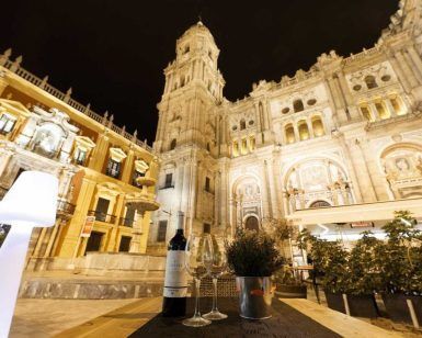 Una vista impresionante de la catedral de Málaga por la noche, complementada con una mesa bellamente puesta del Restaurante L'expérience (Málaga) con una botella de vino y copas, ideal para disfrutar de momentos bajo las estrellas. - ForTwo - Planes para parejas