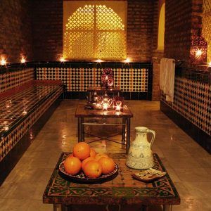 Un acogedor baño marroquí adornado con azulejos ornamentados y velas encendidas presenta el ambiente del Circuito de baños árabes y masaje Boabdil en Aljibe de San Miguel, completo con una jarra decorativa junto a naranjas frescas, que recuerda a los tradicionales baños árabes. - ForTwo - Planes para parejas
