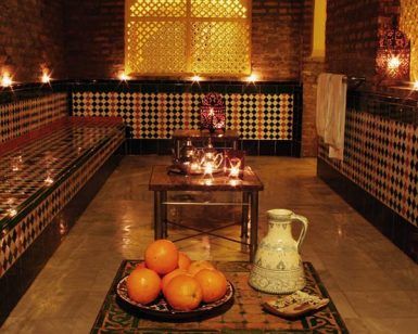 Un acogedor baño marroquí adornado con azulejos ornamentados y velas encendidas presenta el ambiente del Circuito de baños árabes y masaje Boabdil en Aljibe de San Miguel, completo con una jarra decorativa junto a naranjas frescas, que recuerda a los tradicionales baños árabes. - ForTwo - Planes para parejas