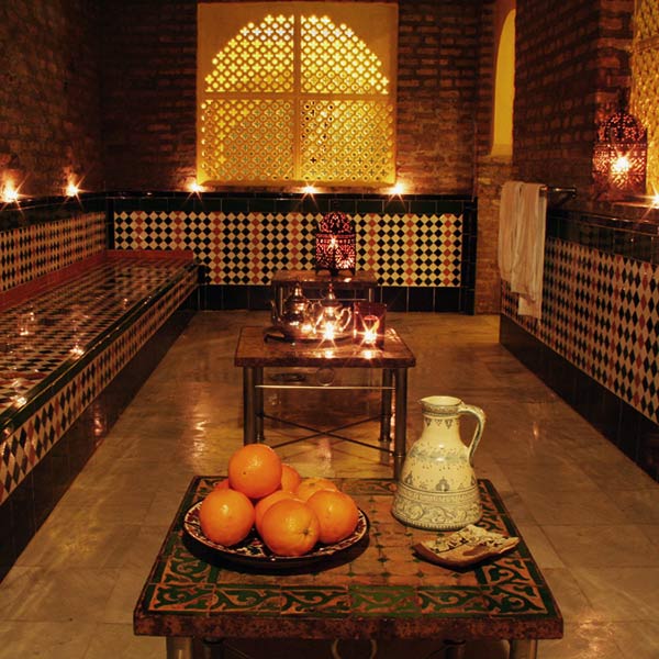 Un acogedor baño marroquí adornado con azulejos ornamentados y velas encendidas presenta el ambiente del Circuito de baños árabes y masaje Boabdil en Aljibe de San Miguel, completo con una jarra decorativa junto a naranjas frescas, que recuerda a los tradicionales baños árabes. - ForTwo - Planes para parejas