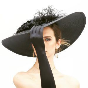 Una mujer con sombrero negro y guantes se cubre la mitad de la cara con la mano contra un fondo de estudio blanco, exudando elegancia y misterio que recuerda a una clásica "Sesión de fotografía de pareja de estudio (Madrid)", combinando el encanto sin esfuerzo. - ForTwo - Planes para parejas