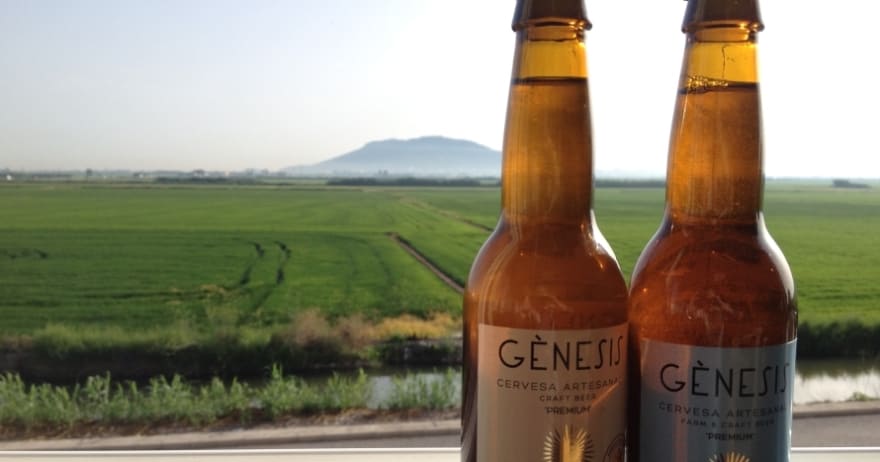 Dos botellas de cerveza se yerguen ante un exuberante campo verde con una montaña a lo lejos bajo un cielo despejado, capturando la esencia de Valencia. Te sentirás como en una "Visita a la fábrica de cerveza Génesis con cata y almuerzo" en Valencia, saboreando la belleza natural que inspira cada cerveza. - ForTwo - Planes para parejas