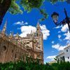 La majestuosa Catedral de Sevilla, con su imponente campanario bajo un cielo azul despejado, está rodeada de frondosos árboles y una encantadora farola. Experimente su belleza atemporal con "Visita la Catedral de Sevilla sin esperas". - ForTwo - Planes para parejas