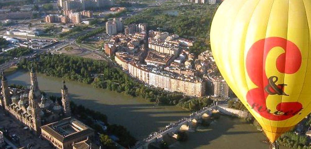 Un globo Vuelo en Globo por Globosreal (Zaragoza) en vibrantes tonos amarillos y rojos se eleva con gracia sobre Zaragoza, resaltando sus edificios históricos y su serpenteante río. - ForTwo - Planes para parejas